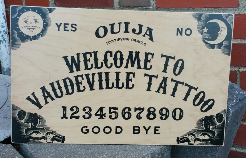 Pu&ograve; includere: Una tavola Ouija in legno con scritte e illustrazioni nere. La tavola dice "Ouija Mystifying Oracle" e "Welcome to Vaudeville Tattoo". Presenta i numeri 0-9, "Yes", "No" e "Good Bye".