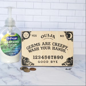 Puede incluir: Una tabla de Ouija de madera con el texto "Germs are creepy. Wash your hands." y los n&uacute;meros 1234567890. La tabla mide 10,16 cm por 15,24 cm y tiene un dise&ntilde;o en blanco y negro.
