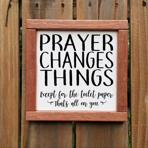 May include: A wooden framed sign with the text "PRAYER CHANGES THINGS Except for the toilet paper That's all on you" in black lettering on a white background.