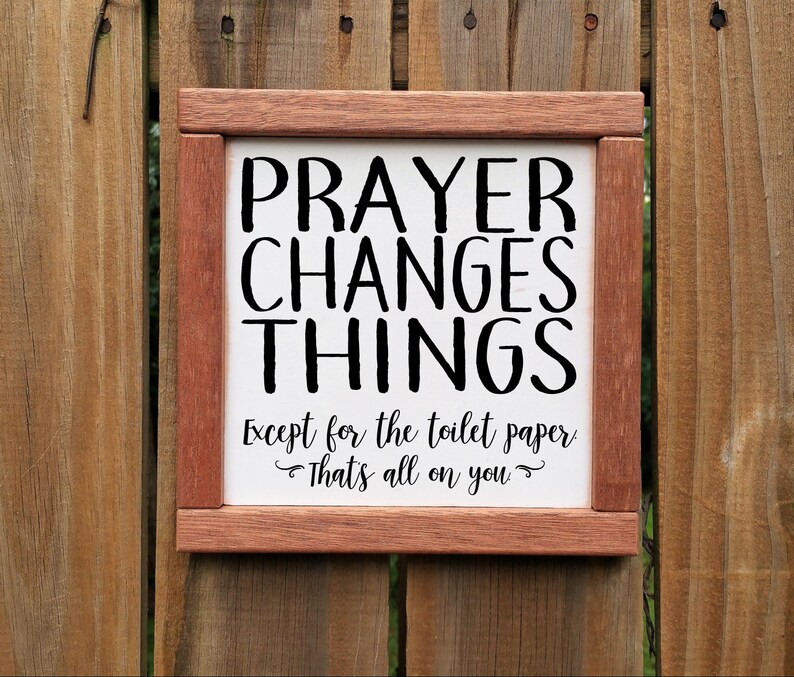 May include: A wooden framed sign with the text "PRAYER CHANGES THINGS Except for the toilet paper That's all on you" in black lettering on a white background.
