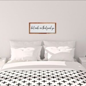 May include: A white wooden sign with the text "don't make me throat punch you" in black cursive lettering. The sign is framed in a light brown wood frame and is hanging on a white wall above a bed with white bedding and a black and white patterned duvet cover.