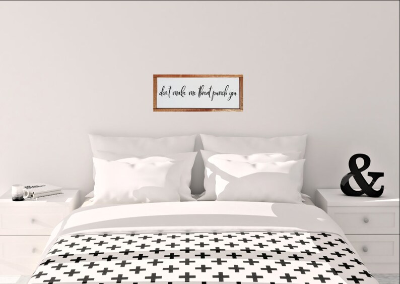 May include: A white wooden sign with the text "don't make me throat punch you" in black cursive lettering. The sign is framed in a light brown wood frame and is hanging on a white wall above a bed with white bedding and a black and white patterned duvet cover.
