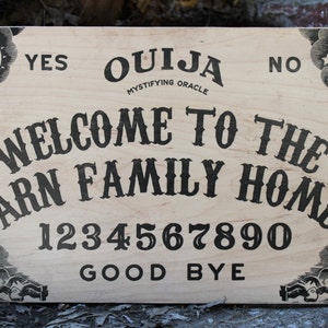 Pu&ograve; includere: Una tavola Ouija in legno con la scritta "Welcome to the Arn Family Home". La tavola presenta le parole "Yes", "No" e "Good Bye", insieme ai numeri 0-9. Grafiche decorative di sole e luna negli angoli.