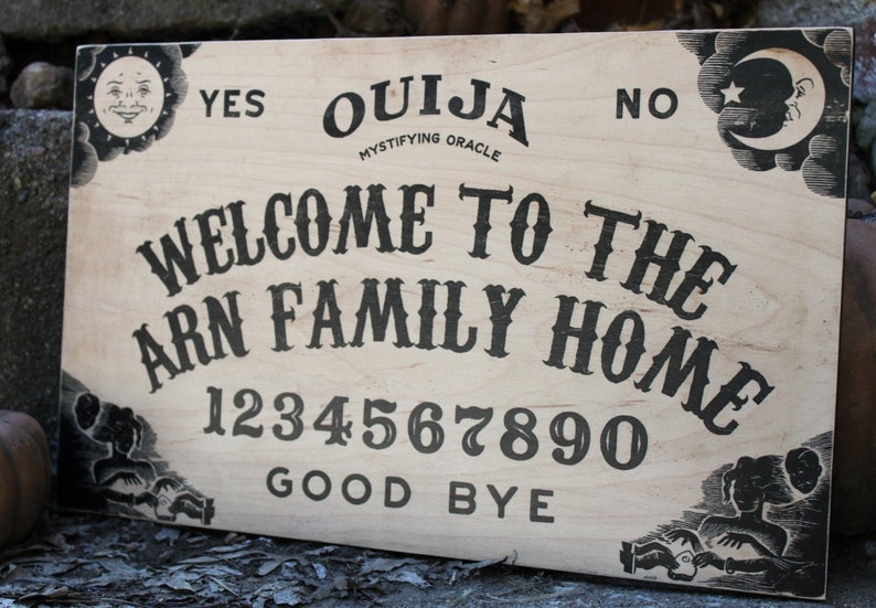 Pu&ograve; includere: Una tavola Ouija in legno con scritte e illustrazioni nere. La tavola presenta le parole "Ouija", "Welcome to the Arn Family Home", numeri e le frasi "Yes", "No" e "Good Bye". Grafiche decorative di sole e luna negli angoli.