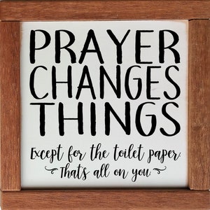 May include: A white wooden sign with black text that reads "Prayer Changes Things Except for the toilet paper. That's all on you." The sign is framed in a brown wood frame.