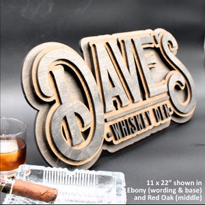 May include: A wooden sign with the text "Dave's Whiskey Den" in a layered, 3D style. The sign is made of ebony and red oak wood and measures 11 inches by 22 inches.