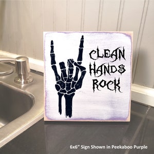 Puede incluir: Un letrero de madera de 15,24 x 15,24 cm con fondo morado. El letrero presenta una mano de esqueleto negra haciendo el gesto de rock and roll. El texto "Clean Hands Rock" est&aacute; escrito en una fuente g&oacute;tica negra.