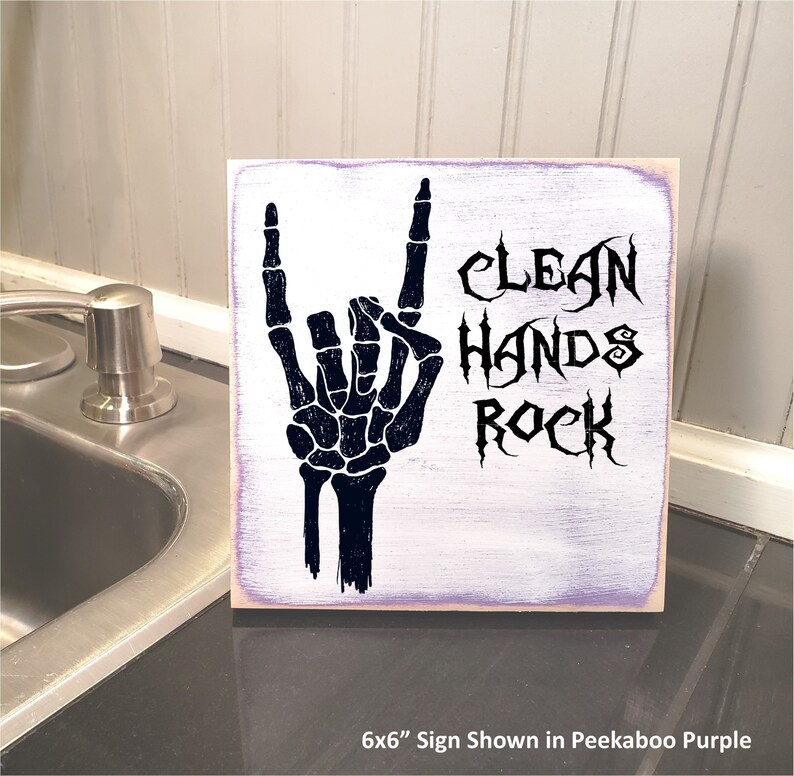 Puede incluir: Un letrero de madera de 15,24 x 15,24 cm con fondo morado. El letrero presenta una mano de esqueleto negra haciendo el gesto de rock and roll. El texto "Clean Hands Rock" est&aacute; escrito en una fuente g&oacute;tica negra.