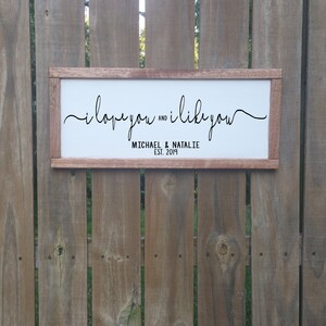 May include: A white wooden sign with the text "i love you and i like you" in black cursive script. Below the text is "MICHAEL & NATALIE EST. 2019" in black.