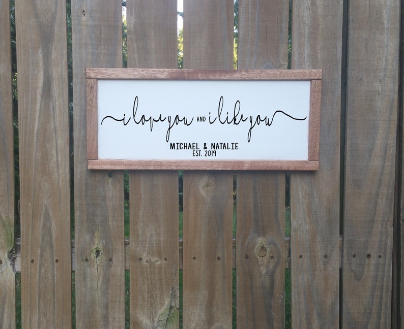 May include: A white wooden sign with the text "i love you and i like you" in black cursive script. Below the text is "MICHAEL & NATALIE EST. 2019" in black.