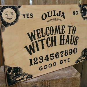 Pu&ograve; includere: Un tabellone Ouija in legno con scritte e illustrazioni nere. Il tabellone presenta le parole "Ouija", "Welcome to Witch Haus", numeri e le frasi "Yes", "No" e "Good Bye". Grafiche decorative di sole e luna negli angoli.