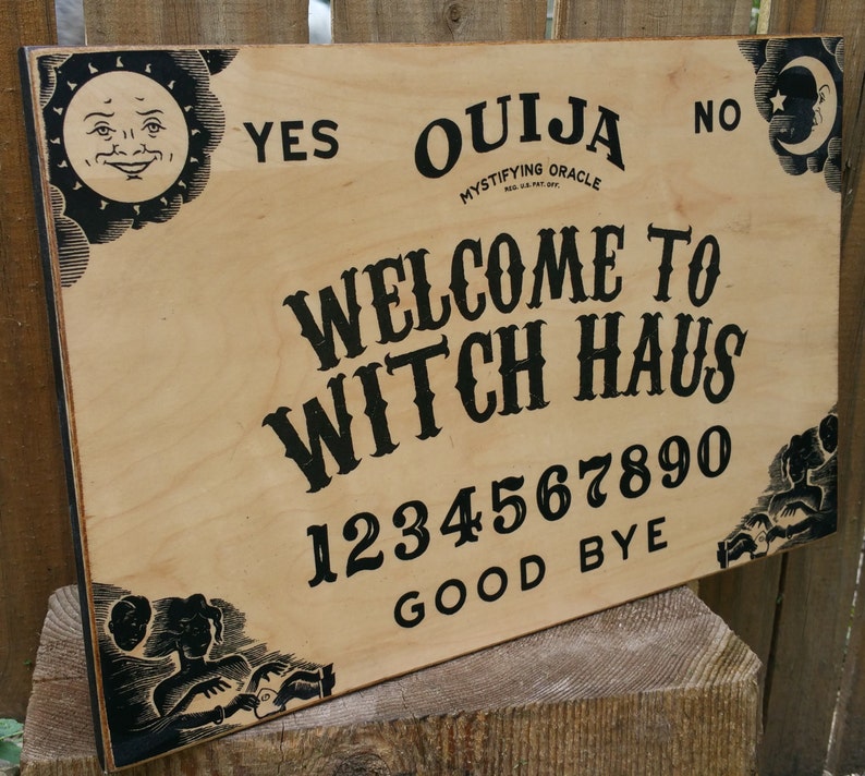 Pu&ograve; includere: Un tabellone Ouija in legno con scritte e illustrazioni nere. Il tabellone presenta le parole "Ouija", "Welcome to Witch Haus", numeri e le frasi "Yes", "No" e "Good Bye". Grafiche decorative di sole e luna negli angoli.