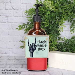 Puede incluir: Un letrero de madera de 7,6 x 7,6 cm con un fondo azul claro y una mano de esqueleto negra haciendo el gesto de rock and roll. El letrero dice "Clean Hands Rock". El letrero est&aacute; colgado de una botella de vidrio marr&oacute;n con una tapa roja.