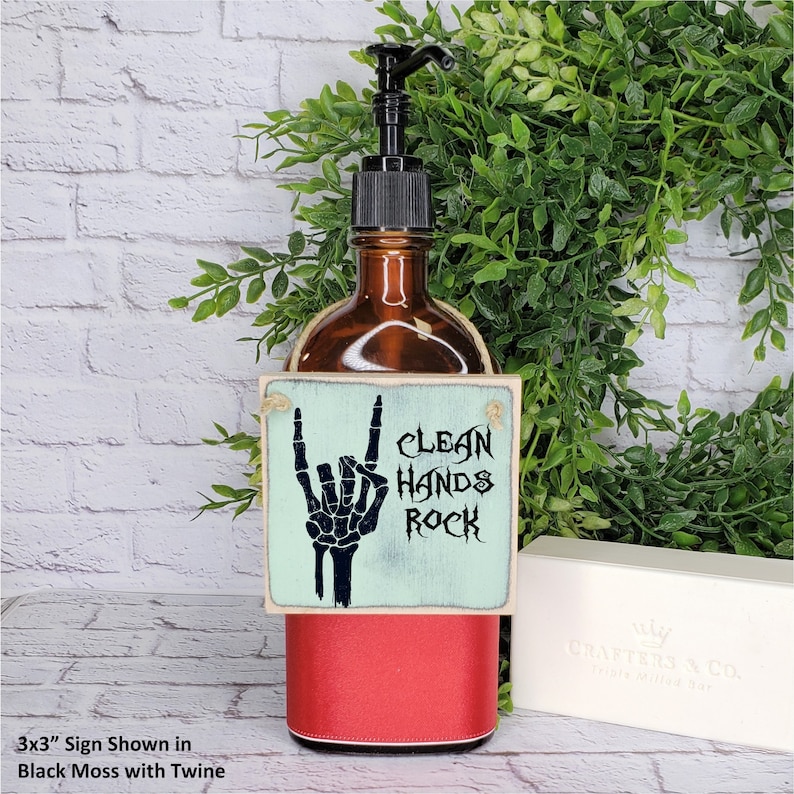 Puede incluir: Un letrero de madera de 7,6 x 7,6 cm con un fondo azul claro y una mano de esqueleto negra haciendo el gesto de rock and roll. El letrero dice "Clean Hands Rock". El letrero est&aacute; colgado de una botella de vidrio marr&oacute;n con una tapa roja.