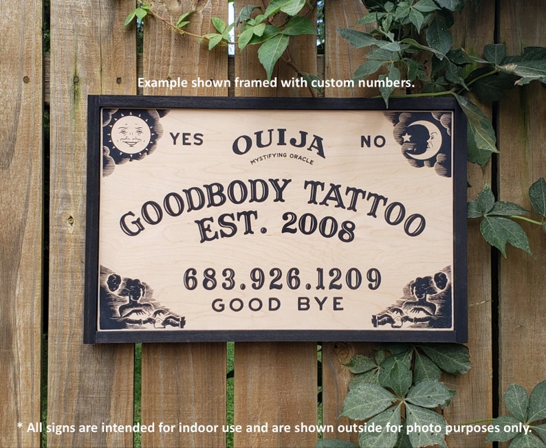 Pu&ograve; includere: Una tavola Ouija in legno con cornice nera. Presenta le parole "YES" e "NO", e "OUIJA MYSTIFYING ORACLE". Mostra anche "GOODBODY TATTOO EST. 2008", un numero di telefono e "GOOD BYE". Gli angoli sono decorati con disegni di sole e luna.