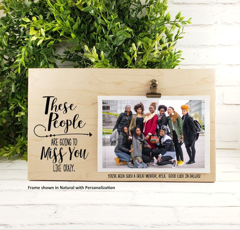 May include: A wooden photo frame with a black inscription that reads "These People Are Going To Miss You Like Crazy." The frame has a clip to hold a photo. The photo shows a group of people standing together. The text below the photo reads "You've Been Such A Great Mentor, Ayla Good Luck In Dallas!"