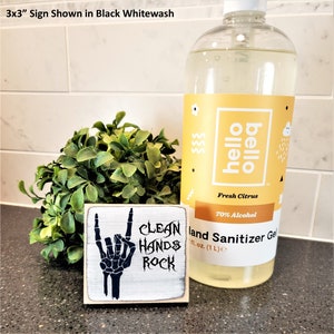 Puede incluir: Un letrero de madera de 7,62 cm x 7,62 cm con un acabado blanqueado y letras negras. El letrero presenta una mano de esqueleto haciendo un gesto de rock and roll con el texto "Clean Hands Rock".