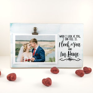 May include: A light blue wooden photo frame with a metal clip holding a photo of a couple. The frame has the text "WHEN I LOOK AT YOU, I CAN FEEL IT. I look at You and I'm Home". Red glitter heart decorations are scattered around the frame.