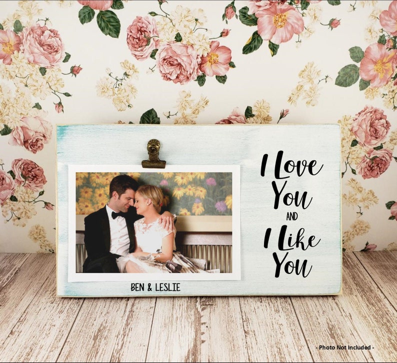K&ouml;nnte beinhalten: Ein wei&szlig;er Holzrahmen mit blauer Waschung und einem Clip oben. Der Rahmen enth&auml;lt ein Foto eines Paares und den Text "I love you and I like you" auf der rechten Seite. Das Foto ist mit "Ben & Leslie" beschriftet.