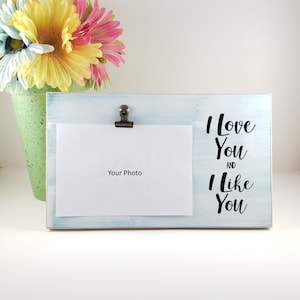 K&ouml;nnte beinhalten: Ein wei&szlig;er Holzbilderrahmen mit blauer Waschung. Der Rahmen hat einen Clip, um ein Foto zu halten. Der Text "I love you and I like you" ist in Schwarz auf den Rahmen gedruckt.