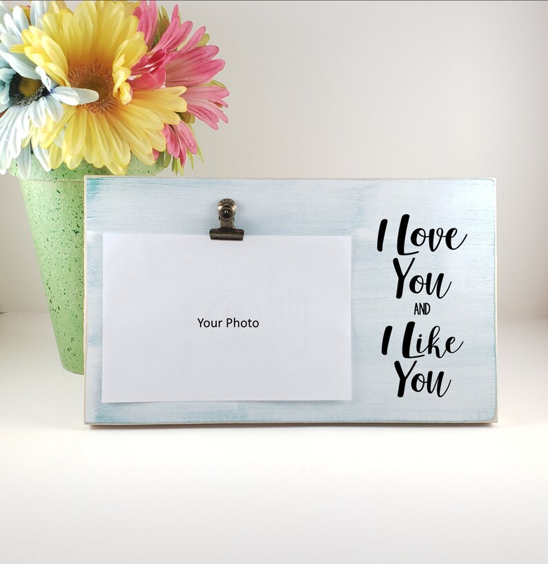 K&ouml;nnte beinhalten: Ein wei&szlig;er Holzbilderrahmen mit blauer Waschung. Der Rahmen hat einen Clip, um ein Foto zu halten. Der Text "I love you and I like you" ist in Schwarz auf den Rahmen gedruckt.