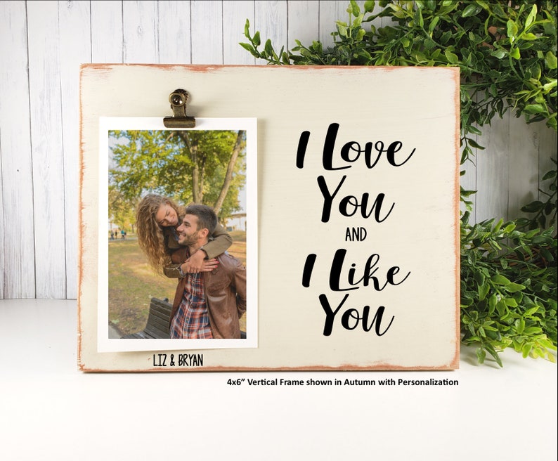 K&ouml;nnte beinhalten: Ein wei&szlig;er Holzrahmen mit einer abgenutzten Oberfl&auml;che. Der Rahmen hat oben einen Clip, um ein Foto zu halten. Das Foto zeigt ein Paar in einem Park. Der Rahmen tr&auml;gt den Text "I Love You And I Like You". Der Rahmen ist 10,2 x 15,2 cm gro&szlig;.