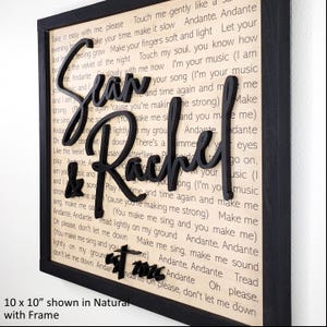 May include: A framed wall art piece featuring the names "Sean & Rachel" in a stylish, black, cursive font. The names are layered over a background of printed song lyrics. The frame is black, and the dimensions are 10 x 10 inches.