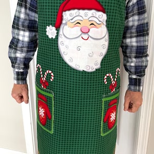May include: Green Christmas apron with a Santa Claus face, candy canes, and mittens. The apron has a checkered pattern and red and white striped straps. The bottom of the apron reads "Happy Holidays!"