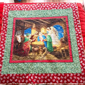May include: A Christmas quilt with a central image of the Nativity scene, framed by a red border with white leaf patterns, a green patterned layer, and a red inner border. The quilt is displayed on a red surface.