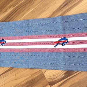 May include: A long, rectangular woven table runner featuring a denim blue base with horizontal red and white stripes running through the center. The design includes repeating Buffalo Bills team logos and finished with blue fringe at both ends.