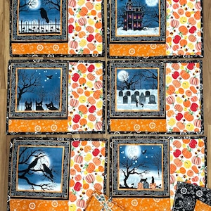 May include: Set of six Halloween-themed placemats with coordinating coasters. Each placemat features a different spooky scene, including a haunted house, graveyard, and black cats, set against a full moon. The borders are black with orange and pumpkin patterns.