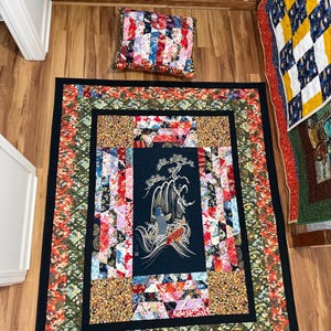 May include: A handmade quilt with a black border, featuring a central panel with a Japanese-style design of a waterfall and koi fish. The quilt incorporates various fabric patterns and colors, including red, green, and gold. A matching pillow is also visible.