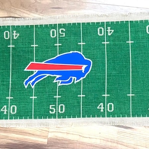 May include: Green burlap table runner with a football field design. Features yard lines, the Buffalo Bills logo, and the words "TOUCHDOWN!" at each end. Includes a brown and white football graphic.