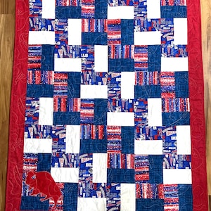 May include: A red, white, and blue patchwork quilt with a Buffalo Bills theme. The quilt is made up of squares of different fabrics, each with a different Bills design. The quilt is edged in red fabric and has a red Buffalo Bills logo in the lower left corner.