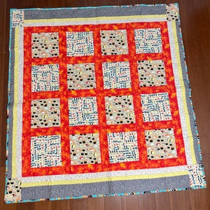 Op de afbeelding: Een quilt met een patchwork-ontwerp met verschillende sushi-rollen. De quilt is gemaakt van verschillende stoffen in tinten oranje, geel en blauw. De quilt is afgewerkt met een grijze en witte bies.