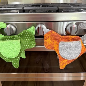 Op de afbeelding: Twee stoffen ovenwanten in de vorm van katten, hangend aan de knoppen van een roestvrijstalen oven. De ene want is groen met witte stippen en een zak met krulpatroon, de andere is oranje met een grijze zak met krulpatroon en een staart.