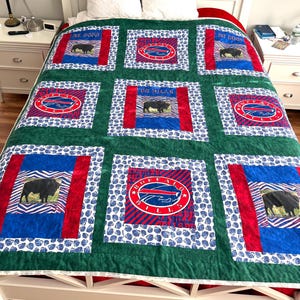 May include: A green and white quilt featuring the Buffalo Bills logo and a buffalo image. The quilt has a patchwork design with red, blue, and white accents. The quilt is on a white bed frame.