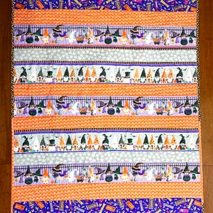Può includere: Una trapunta a tema Halloween con un motivo ripetitivo di gnomi che indossano cappelli da strega e tengono zucche. La trapunta è realizzata in tessuto arancione, viola e grigio con un bordo nero e bianco.