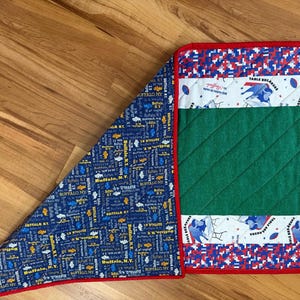 May include: A quilted table runner with a red border. The runner features a green center, white bands with football graphics, and blue and red pixelated patterns. A blue and white patterned fabric is visible on the left.