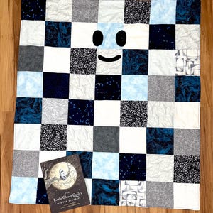 May include: A handmade quilt with a smiling ghost face design. The quilt features a patchwork pattern in shades of blue, white, and gray. A book titled "The Little Ghost Quilt's Winter Surprise" is placed on the quilt.