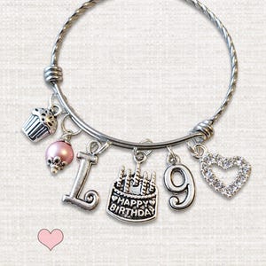 May include: A silver-toned wire bangle bracelet featuring five charms: a cupcake, a pink pearl bead, the letter L, a cake charm with the text Happy Birthday, the number 9, and a heart-shaped charm encrusted with clear crystals.