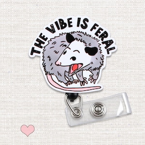 May include: A badge reel featuring a cartoon opossum with gray fur, black ears, and a pink tail. The opossum has an open mouth showing teeth. Above the opossum, the text reads THE VIBE IS FERAL in black capital letters. A clear plastic strap with a metal snap is attached.