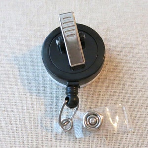 May include: A black retractable badge reel with a silver clip and a black and white design. The reel has a silver ring and a clear plastic badge holder.