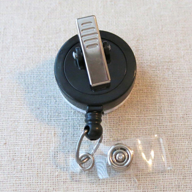 May include: A black retractable badge reel with a silver clip and a black and white design. The reel has a silver ring and a clear plastic badge holder.