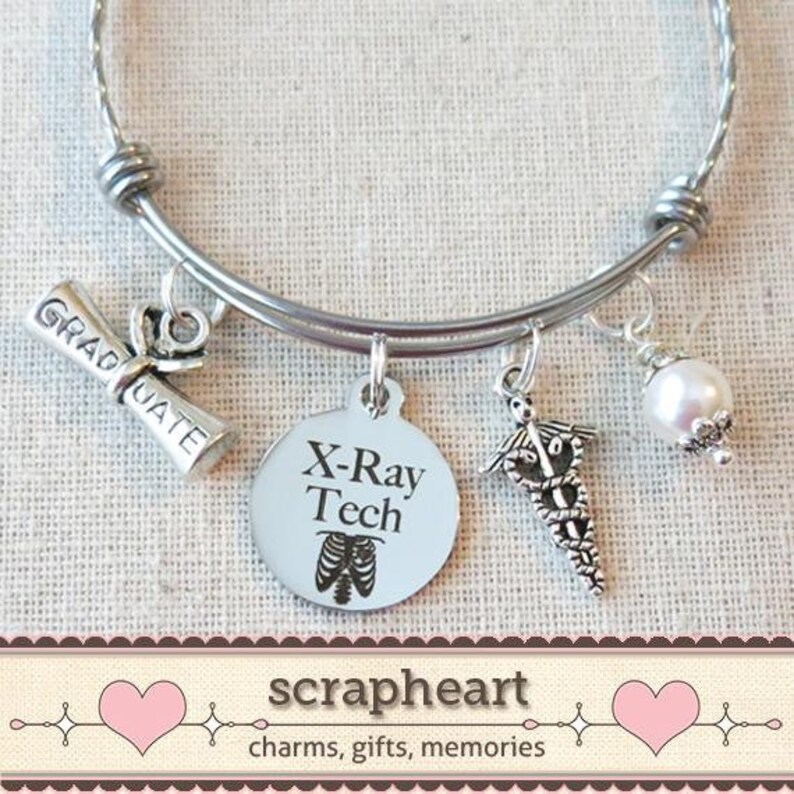 May include: Silver charm bracelet with a variety of charms, including a graduation cap, a charm with the text "X-Ray Tech", a medical symbol charm, and a white pearl charm. The bracelet is on a white fabric background.