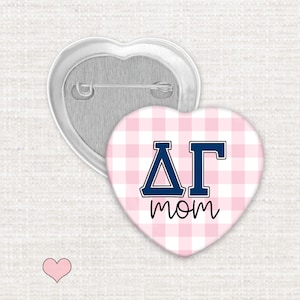 May include: A heart-shaped pinback button featuring a light pink and white gingham pattern. The center displays the Greek letters Delta Gamma in navy blue with a white outline, positioned above the word mom in a black cursive script font.