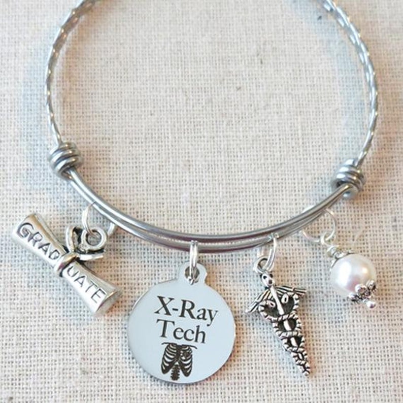May include: Silver bangle bracelet with charms, including a graduation cap, a round charm with the text "X-Ray Tech" and a skeletal image of a ribcage, a caduceus charm, and a white pearl charm.