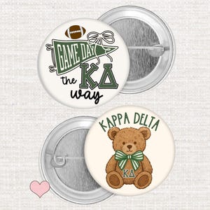 May include: Two round pinback buttons with metal backs. One button features a green pennant, a brown football, and the text "GAME DAY the KΔ way". The second button displays a brown teddy bear wearing a green and white striped bow with "KAPPA DELTA" above.
