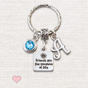 May include: A silver-toned metal keychain featuring a round split ring, a circular charm with a bright blue crystal, a square charm engraved with a sun icon and the text "friends are the sunshine of life", and a decorative letter A charm.