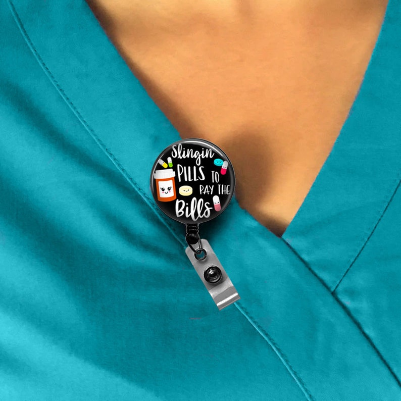 May include: A black and white badge reel with a round button featuring a cartoon pill bottle with a smiling face and the text "Slingin' Pills to Pay the Bills".